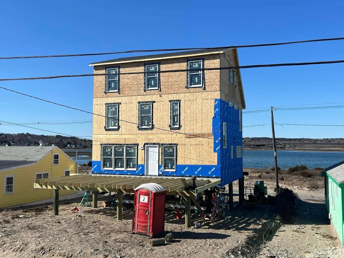 Three-story coastal modular home early in construction — driven-pile stilts foundation, water visible behind, South Shore MA