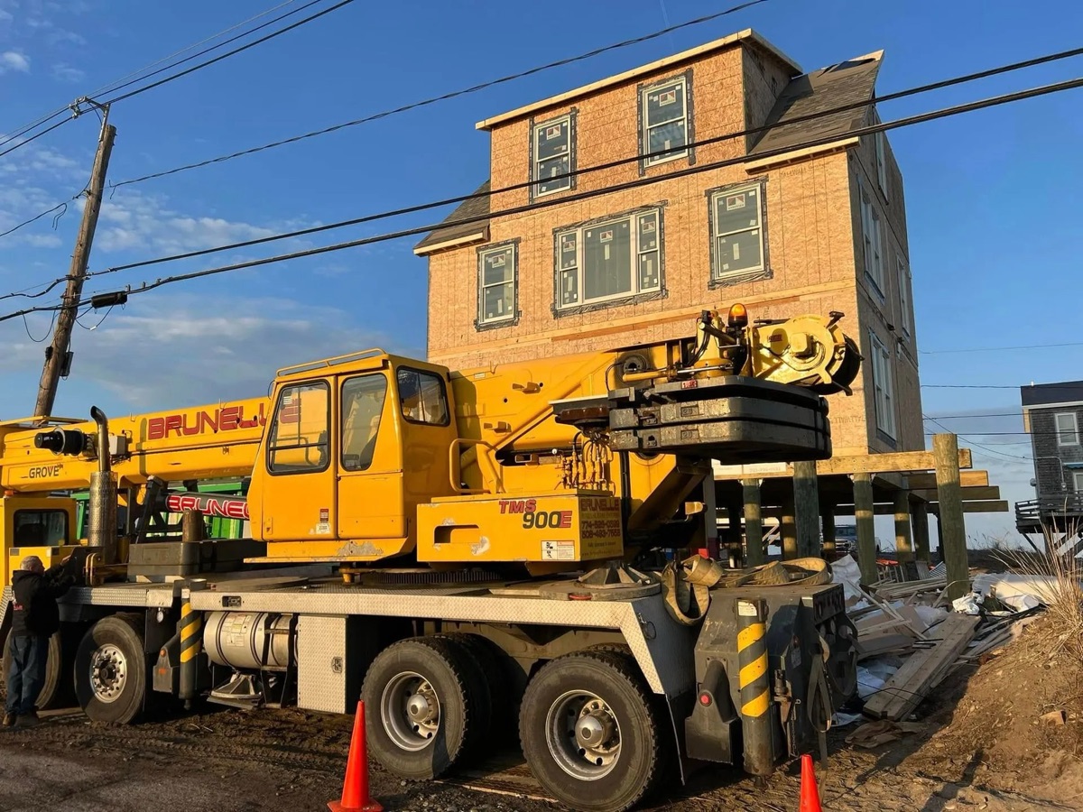 Yellow crane truck on jobsite with a completed three-story coastal modular home in the background