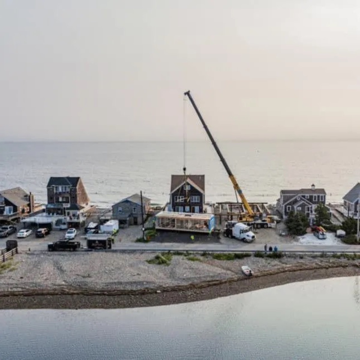Aerial drone view of crane setting a custom modular home on a coastal lot at sunset — South Shore, MA